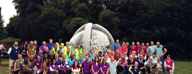 Large group of people wearing colourful bibs posing in front of an inflatable transparent cylinder, imitating the 'crystal maze' from TV.