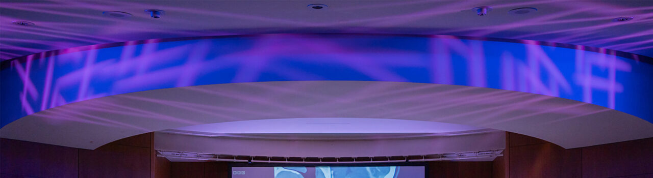 Blue light with pink criss-cross shapes, projected onto ceiling of auditorium