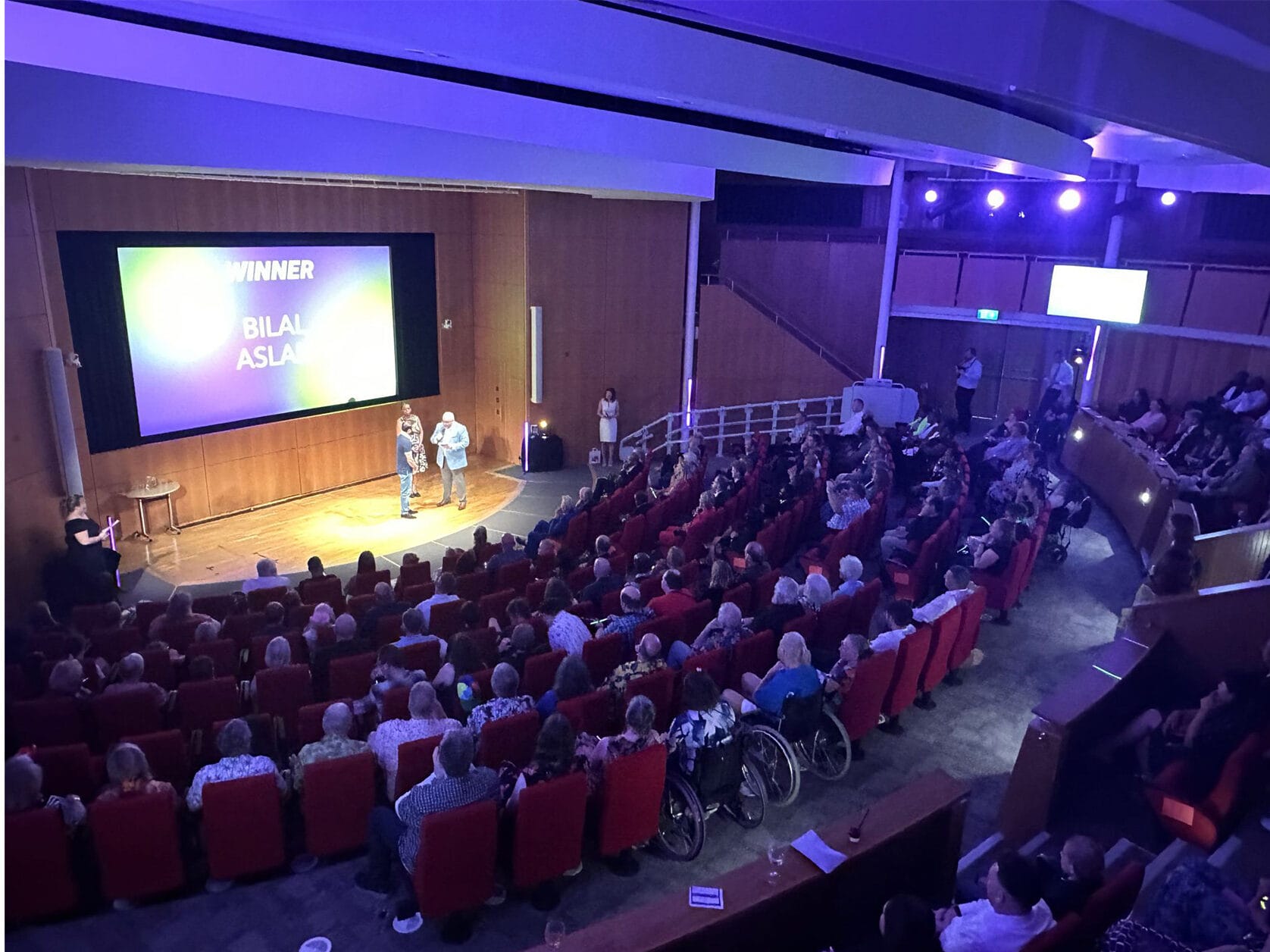 Shot of the Francis Crick Auditorium from the back, showing may seated people including a few in wheelchairs. People watching Bilal Aslam receive his Great Neighbour award.