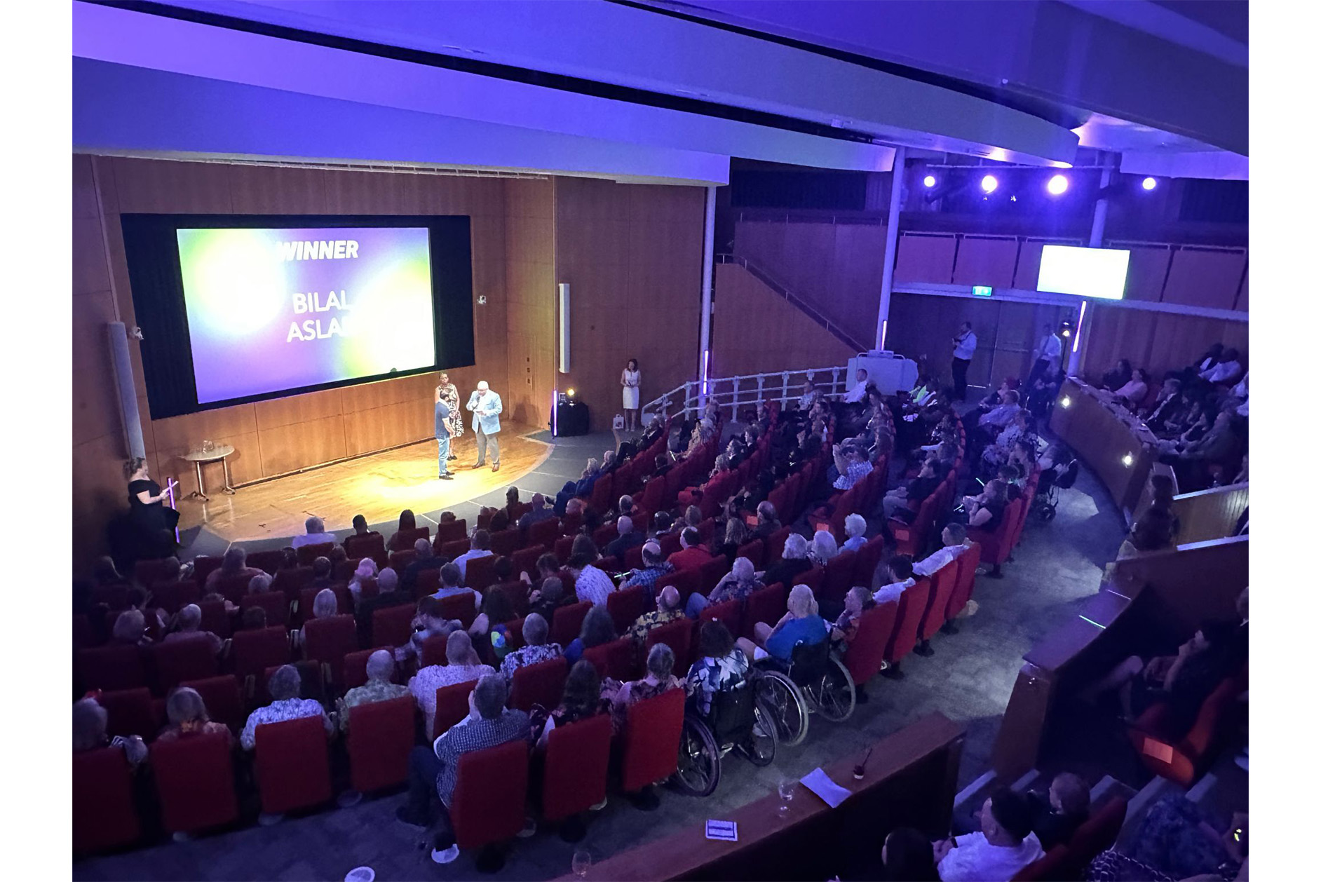 Shot of the Francis Crick Auditorium from the back, showing may seated people including a few in wheelchairs. People watching Bilal Aslam receive his Great Neighbour award.