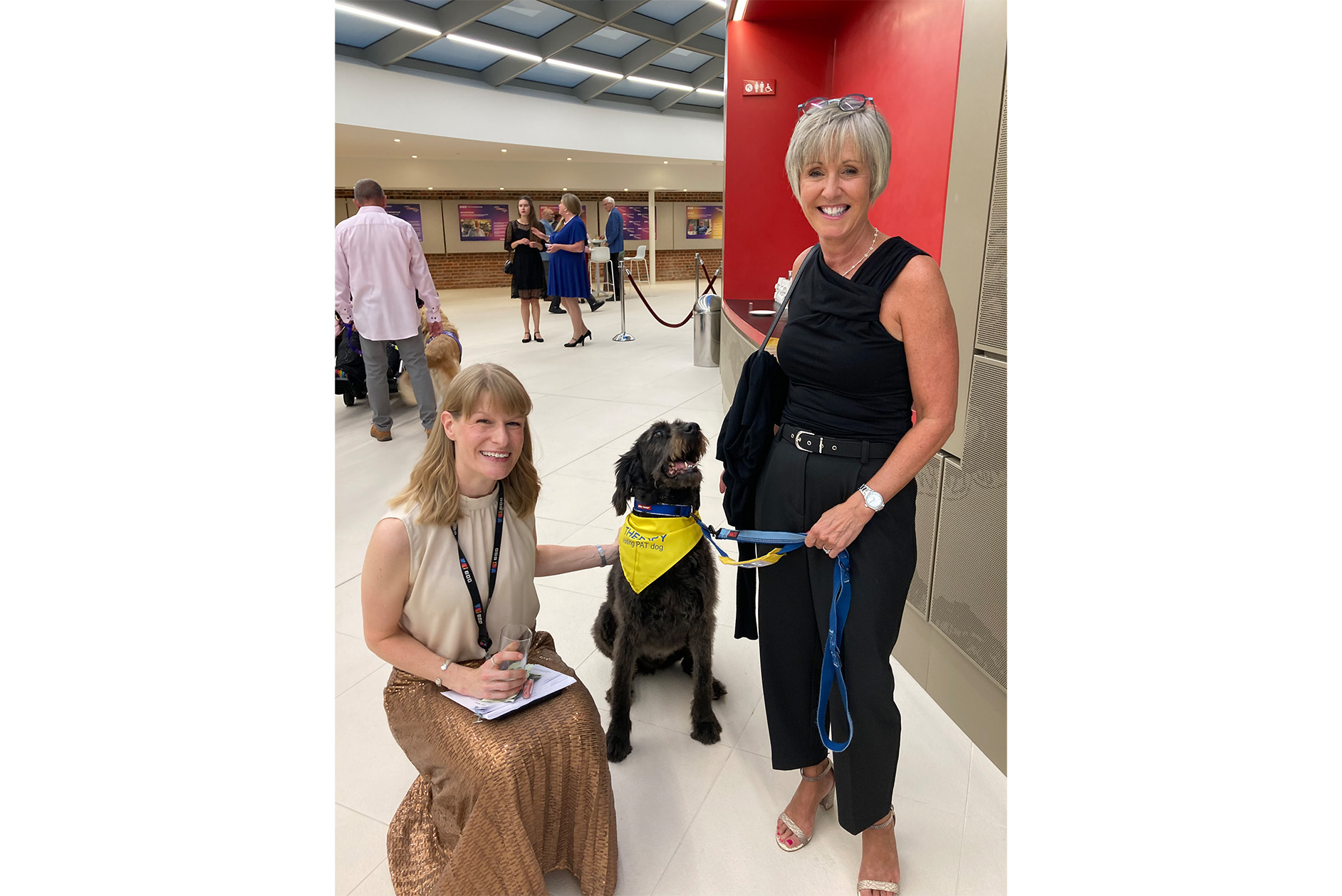 Two women, smiling, on either side of a black labradoodle dog who is wearing a yellow bib that says 'Therapy dog'.