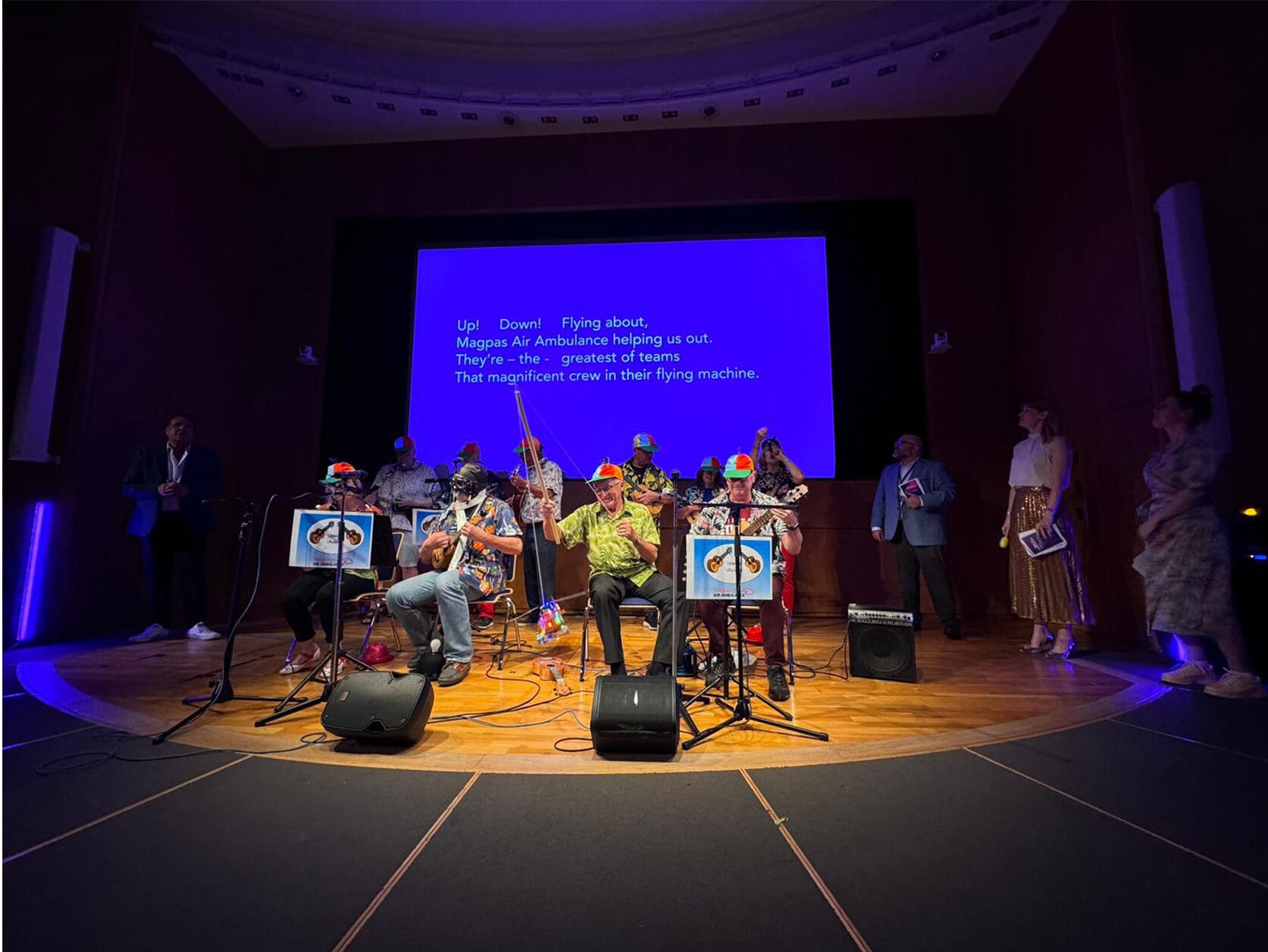 The Upwood Ukuleles group performing on a spot lit stage. Four seated members, 6 standing behind. The words they are singing are projected on the screen behind.