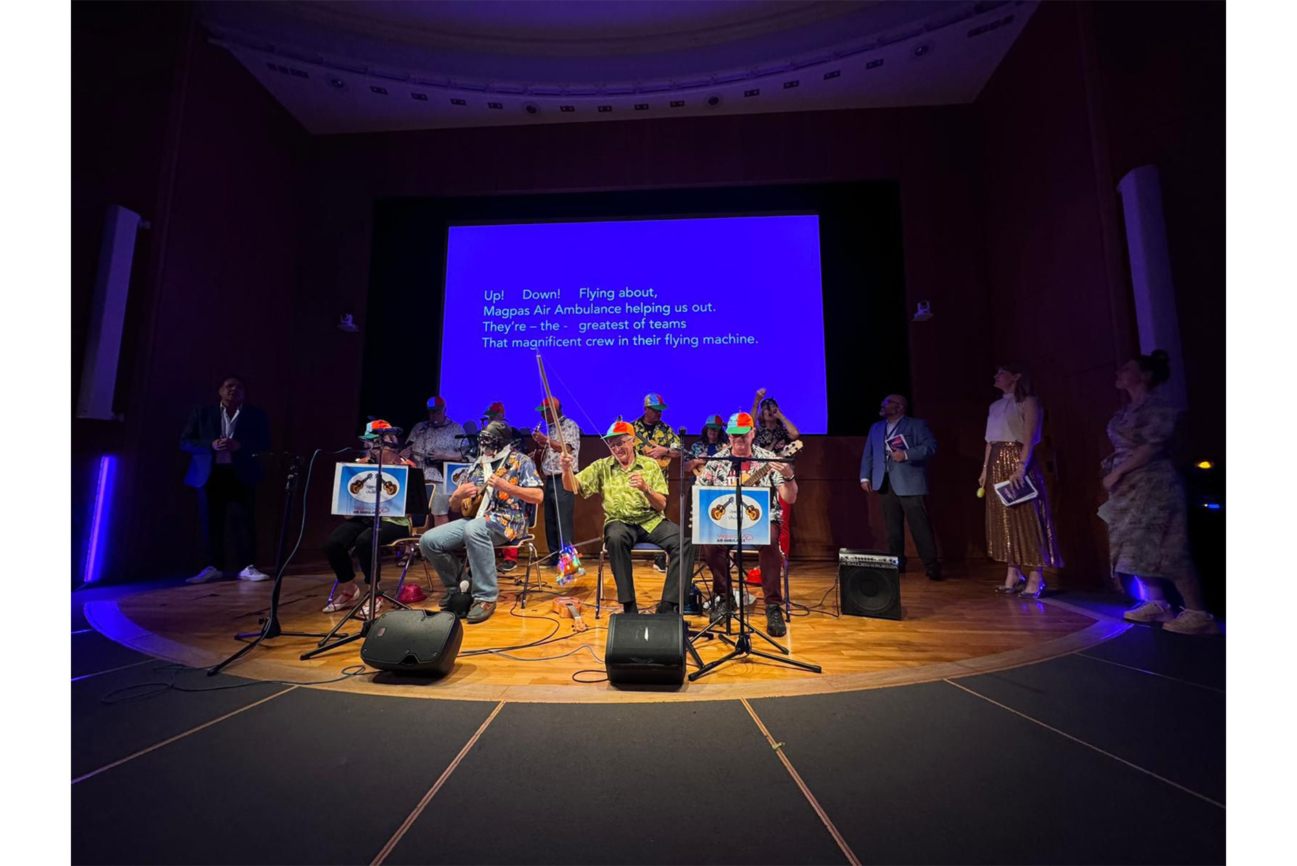 The Upwood Ukuleles group performing on a spot lit stage. Four seated members, 6 standing behind. The words they are singing are projected on the screen behind.