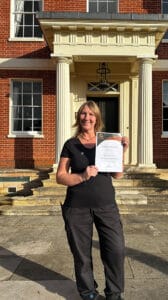 Photo of Hayley Scott standing in the sunshine outside Hinxton Hall holding a piece of paper that's her certificate.