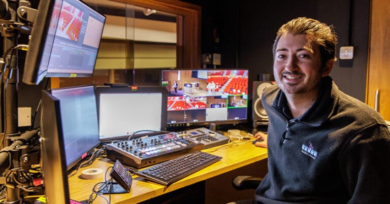 Smiling AV technician sitting at his desk in control booth with screens in front of him and window into auditorium.